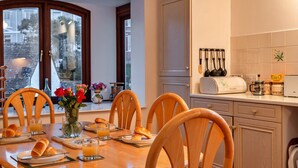 Un coin repas avec une table en bois mise pour le petit-déjeuner, comprenant du pain, des viennoiseries et du jus d’orange.