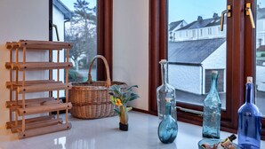 Un présentoir en bois, un panier en osier et des bouteilles en verre sont posés sur une table près d’une fenêtre donnant sur des maisons.
