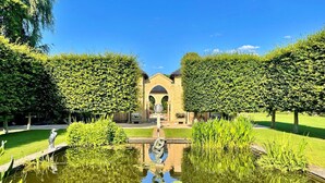 Un jardín tranquilo con un estanque central, un puente de piedra y un edificio con una entrada arqueada.