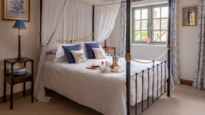 Una cama con dosel y cuatro columnas, ropa de cama blanca y almohadas azules, una mesita de noche con lámpara y una ventana con cortinas.