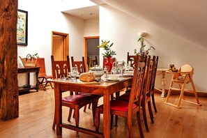 A dining area with a wooden table set for four, surrounded by chairs. There is a wooden chair with a tray, a basket, and a vase with flowers on the table. - The Loft (Kingham)