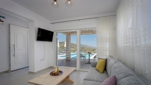 Una sala de estar moderna con sofá, mesa y televisor. Posee un ventanal corredizo que da a un balcón con piscina y montañas al fondo.