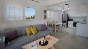 Una sala moderna con un sofá gris, una mesa de centro de madera, y un comedor con una mesa y sillas blancas. Se ve una cocina con armarios y electrodomésticos blancos al fondo.