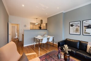 A modern living room with a black leather sofa, a dining area with a white table and chairs, and a kitchen in the background.