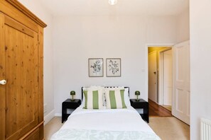 A bedroom with a wooden wardrobe, a bed with green and white striped pillows, two bedside tables with lamps, and framed botanical artwork on the wall.