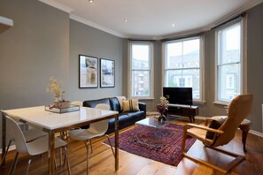 A modern living room with a black leather sofa, a glass coffee table, and a red patterned rug. There is a dining table with chairs and a TV on a stand. - The West Hampstead Place - Inviting 2bdr Apartment (London)