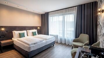 A modern hotel room with a large bed, a bedside table, a chair, a desk with a coffee maker, and a view of the outdoors through sheer curtains.