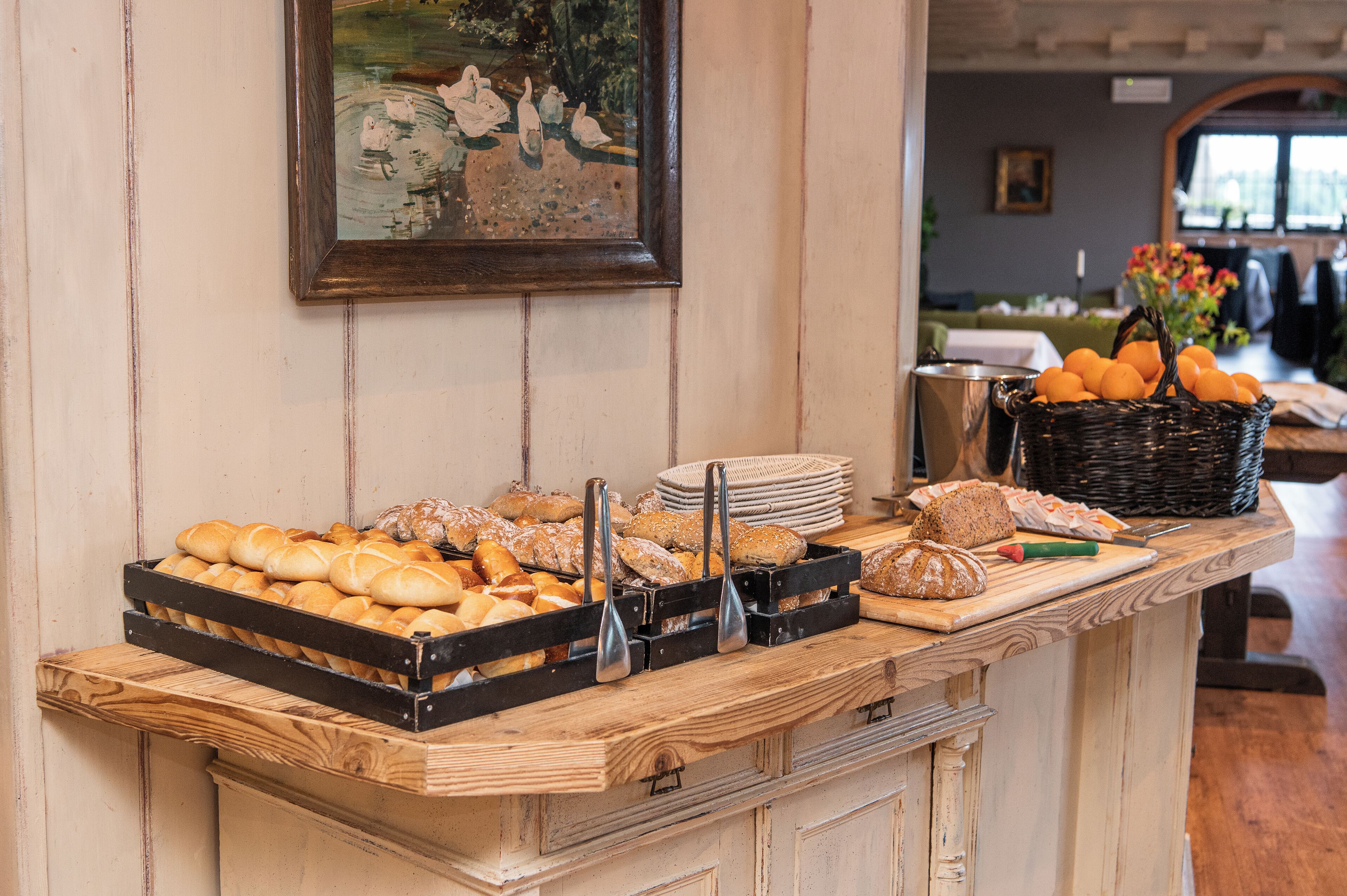 A buffet counter with various breads and pastries, a basket of oranges, and a painting on the wall.