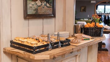 A buffet counter with various breads and pastries, a basket of oranges, and a painting on the wall.