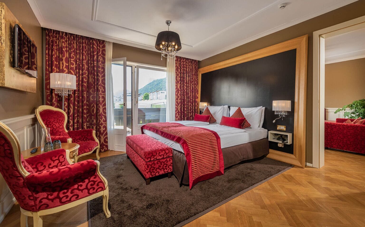 A hotel room with a large bed, red and white bedding, a chandelier, and a view of a mountain.