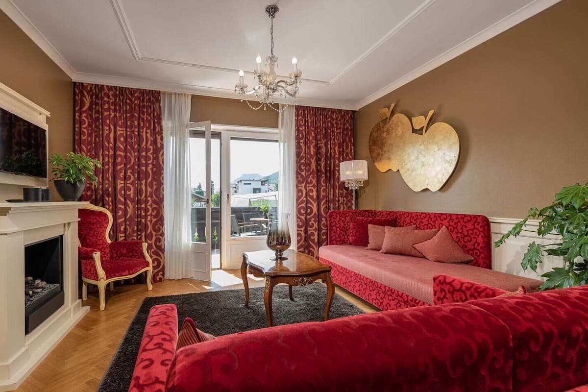 A room with a fireplace, a red sofa, a red armchair, a chandelier, and a large wall decoration.