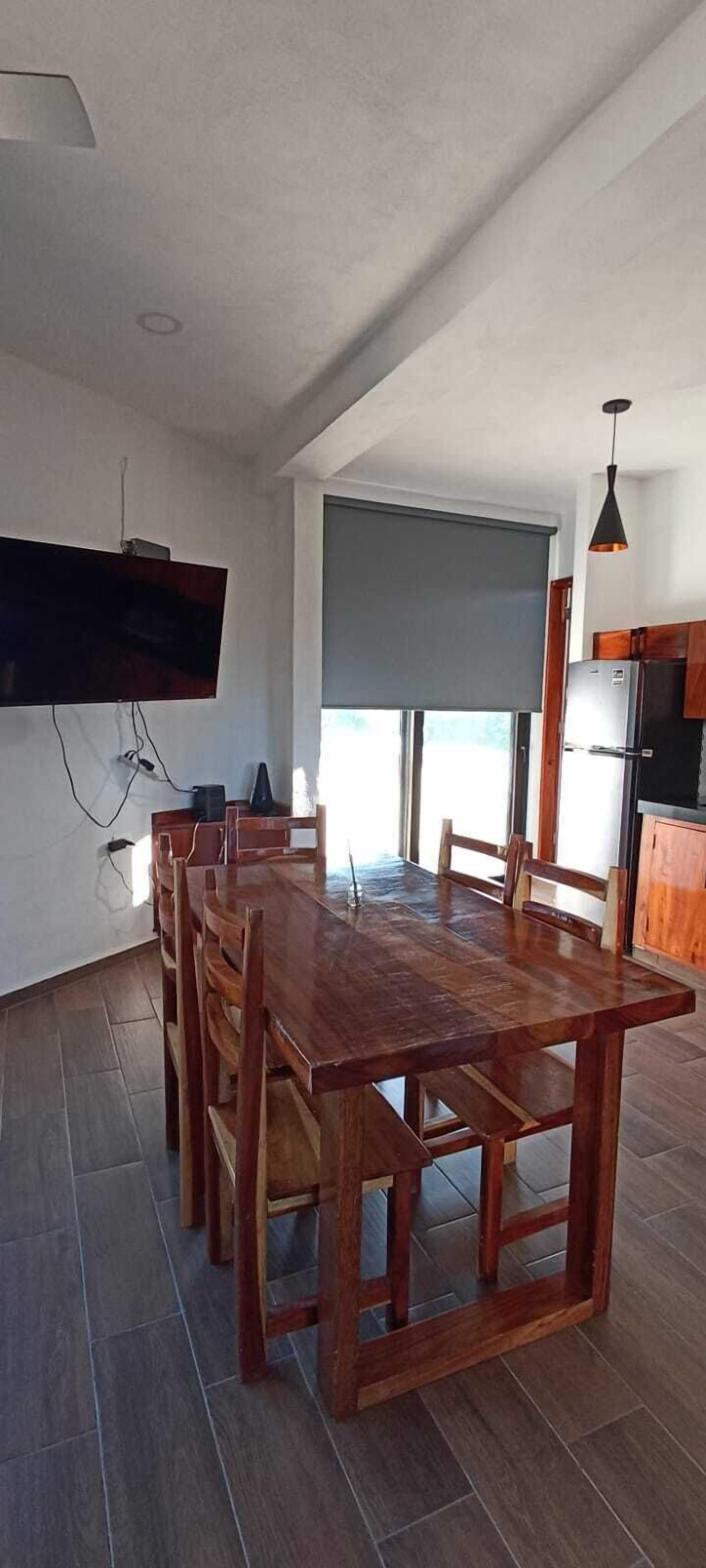 A spacious dining area with a long wooden table and chairs, a flat-screen TV mounted on the wall, and a view of the outdoors through a large window.