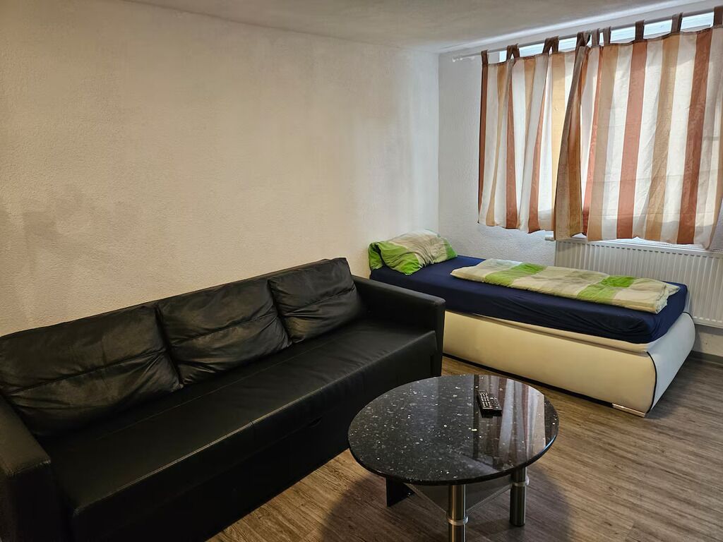 A room with a black leather sofa, a small round table, a bed with a blue mattress and green pillows, and a window with striped curtains.