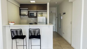Una cocina moderna con encimera blanca, dos taburetes negros, un microondas, un refrigerador y una planta en maceta.