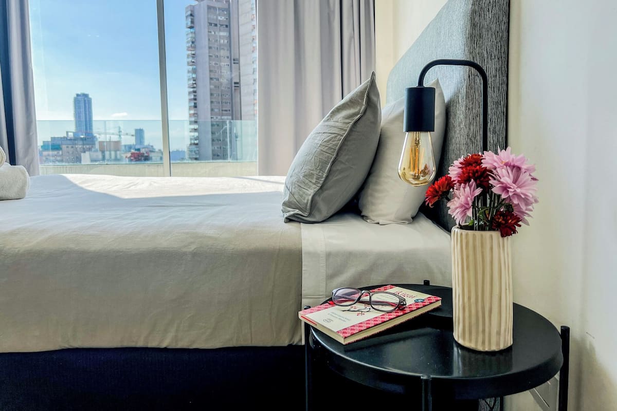 A hotel room with a bed, a bedside table with a book and glasses, a vase with flowers, a lamp, and a city view.