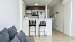 Un salón moderno con un sofá gris y dos sillas negras. En el fondo, se ve una cocina con un microondas y un refrigerador blanco.