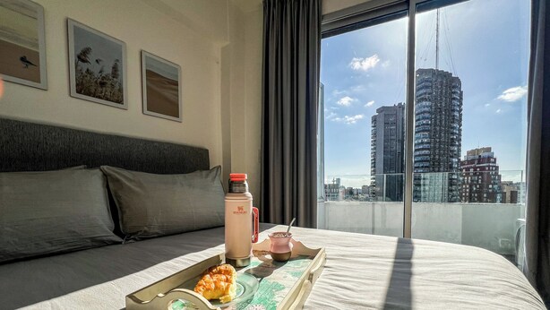 Habitación de hotel con una cama grande, una bandeja con croissant y una bebida, y vista a un paisaje urbano a través de la ventana.
