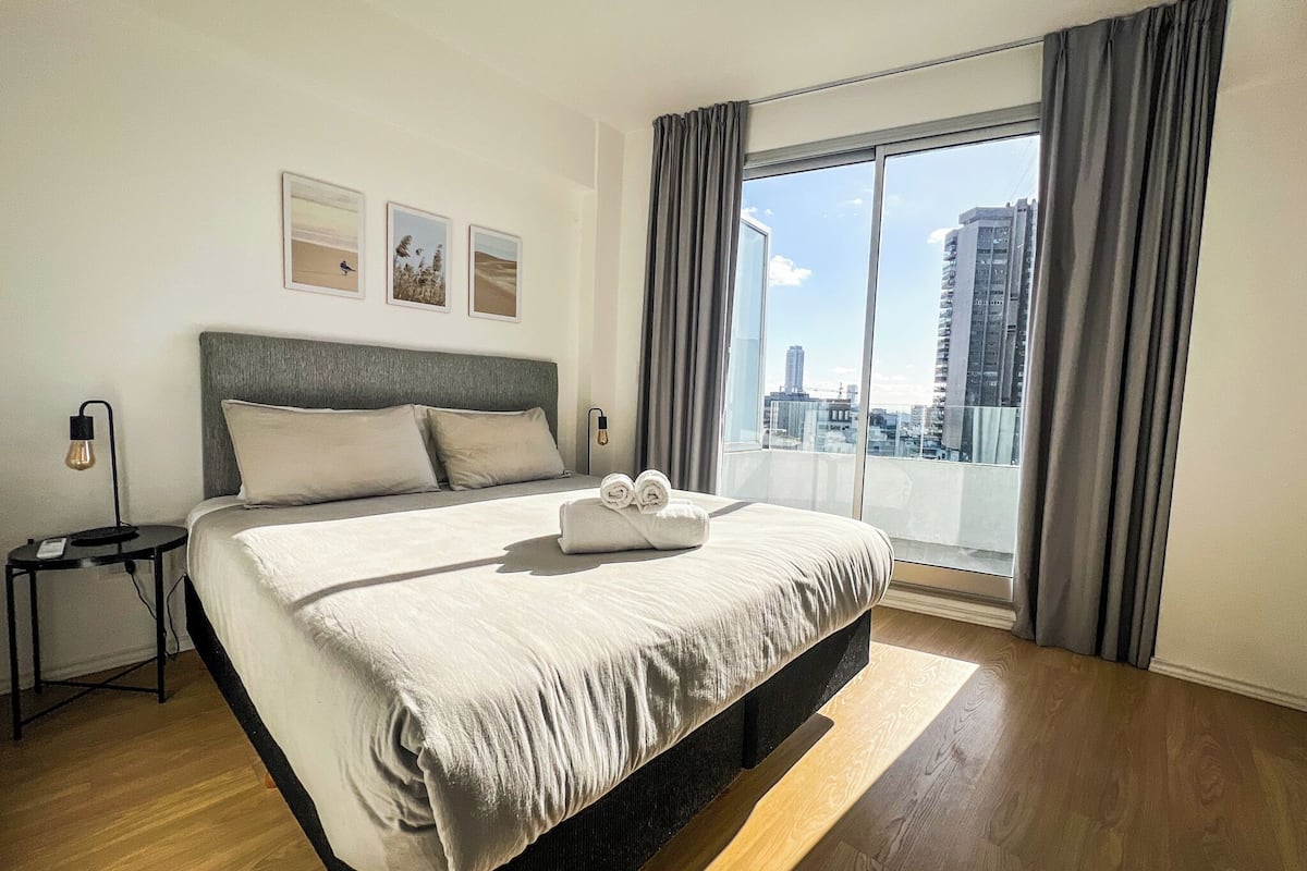 A modern hotel room with a large bed, a nightstand, a lamp, and a city view through the window.