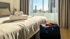Una habitación de hotel con una cama, una maleta y vistas a un paisaje urbano a través de amplias ventanas.