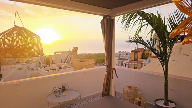 Un balcon avec vue sur le coucher de soleil, un luminaire suspendu, une petite table et une plante en pot.