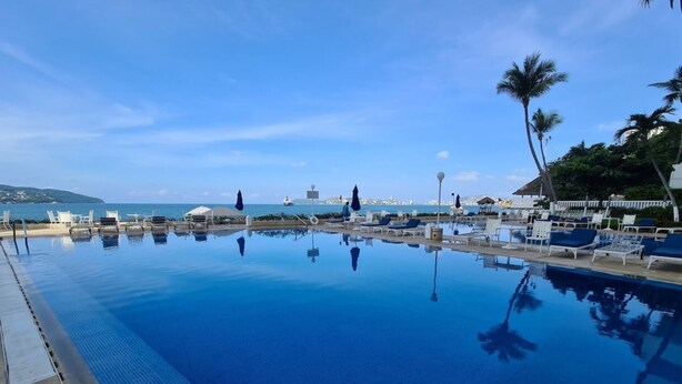 A clear blue infinity pool with lounge chairs and umbrellas, overlooking a coastal area with palm trees and a distant view of a lighthouse.