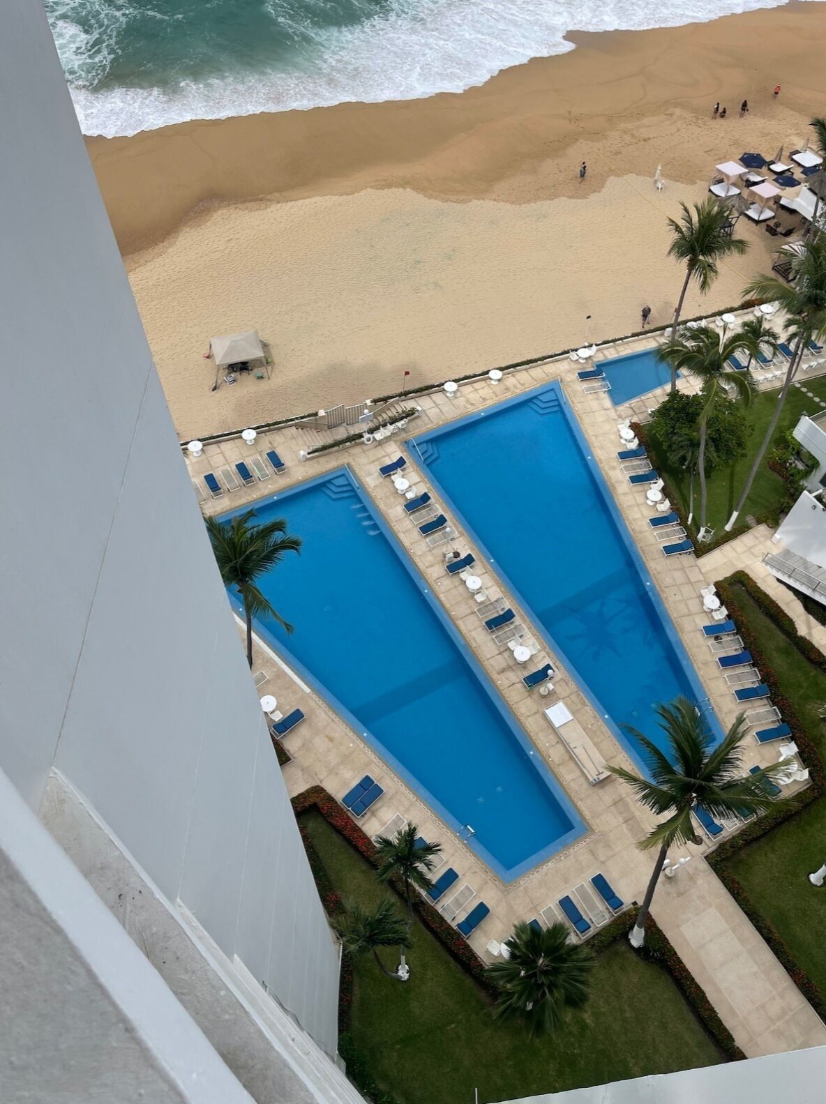 Un espace piscine comprenant deux grandes piscines, entouré de chaises longues et de palmiers, et donnant directement sur une plage de sable.