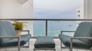 A balcony with two striped armchairs and a small ottoman, overlooking a body of water and a distant shoreline.