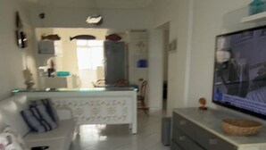 Sala de estar com sofá branco, TV em suporte e cozinha ao fundo.