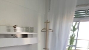 Quarto com cama, parede branca, janela com cortinas e planta lá fora.