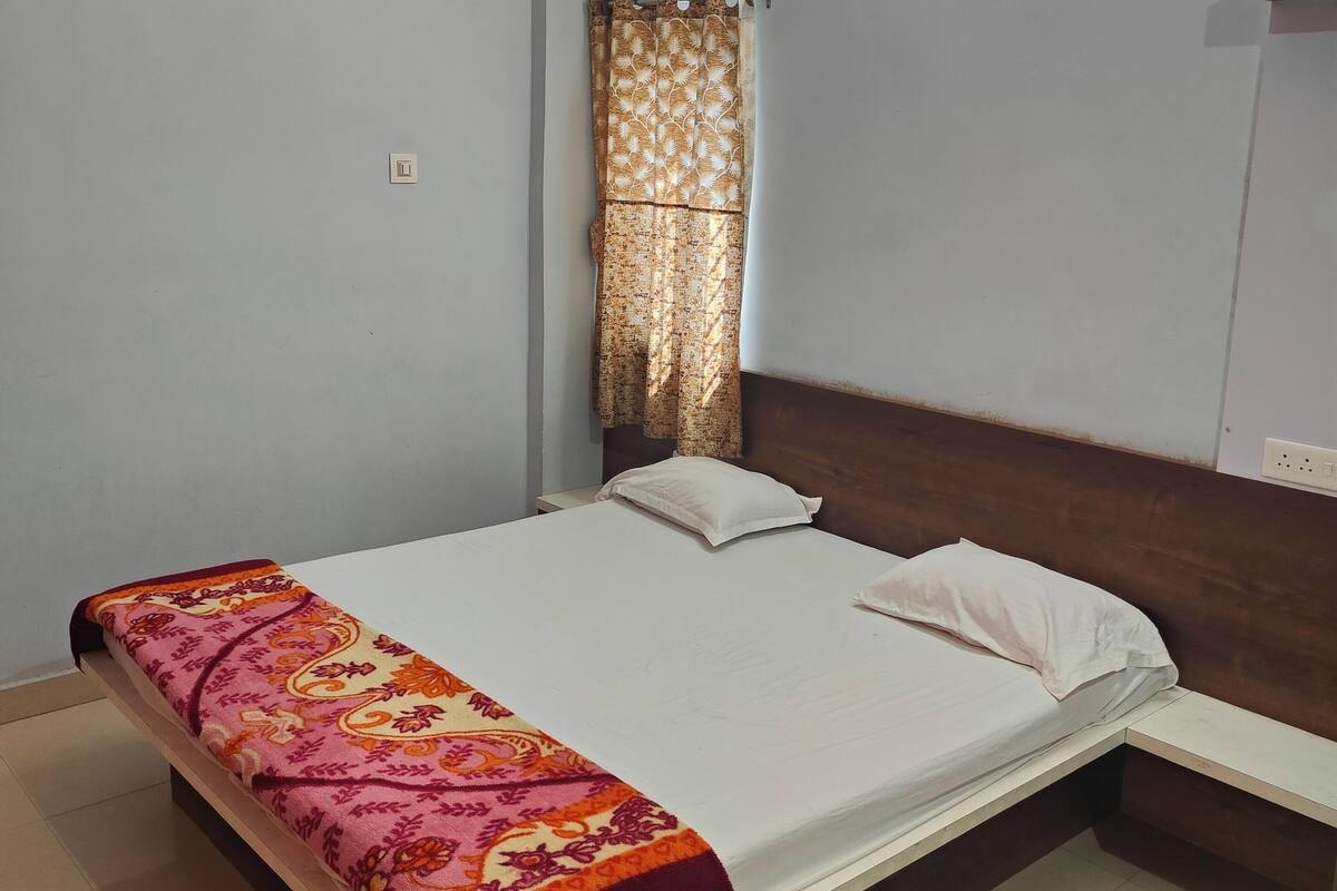 A hotel room with a bed, a bedside table, a nightstand, a wall-mounted air conditioner, a ceiling light, and a curtain.