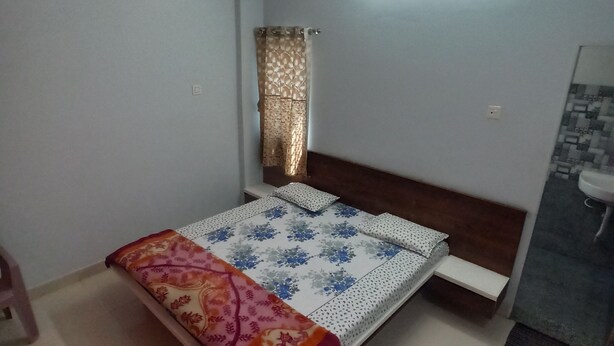 Ein Schlafzimmer mit Bett, Nachttisch, Fenster mit Vorhängen, einer Wandleuchte und einem Badezimmer mit Waschbecken und Spiegel.