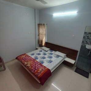 A bedroom with a bed, a nightstand, a window with curtains, a wall-mounted light, and a bathroom with a sink and mirror. - Hotel adiraj palace and lodging (Nevasa)