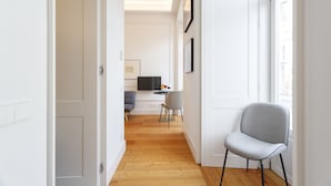 Un couloir moderne et bien éclairé, doté d’un plancher en bois, mène à un coin repas où se trouvent une chaise grise et une petite table.