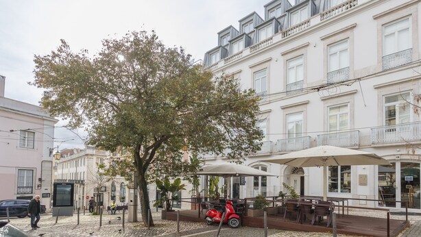 Une rue pavée, avec un arbre, un scooter rouge et un café doté d’une terrasse.