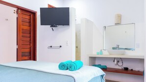 Un dormitorio con cama, televisor montado en la pared, baño con lavabo y espejo, y una puerta de madera.