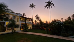 Un edificio moderno de varios pisos con un jardín bien cuidado, palmeras y un cielo despejado al atardecer.