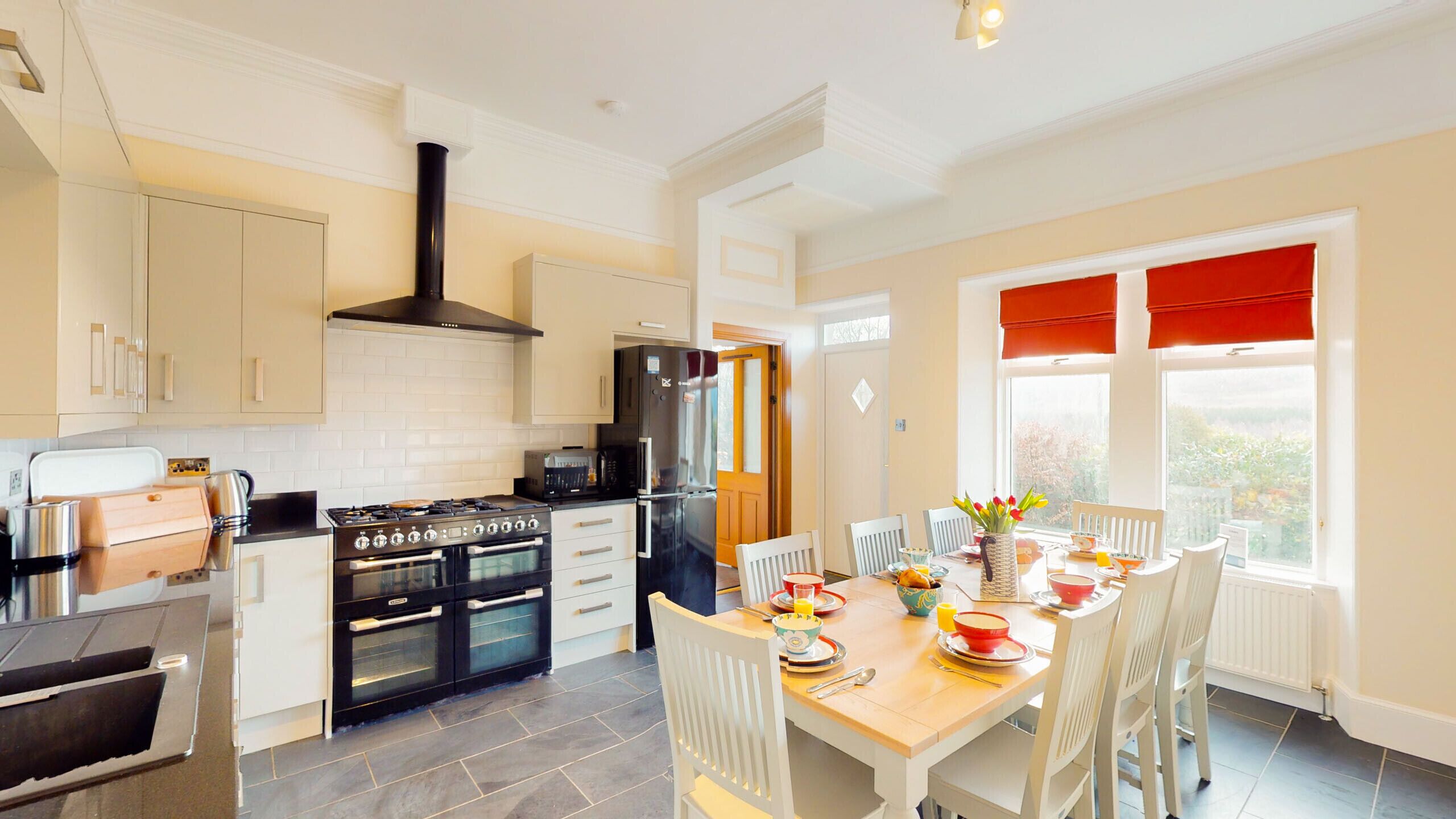 A modern kitchen with a stove, oven, and refrigerator. There is a dining table with chairs and a window with red curtains.