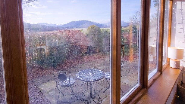 Vista desde una ventana que da a un patio con una mesa y sillas, una lámpara y un paisaje con árboles y montañas.