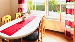 A dining area with a table set for four, chairs, a window with curtains, and a radiator. - Ferienhof Eckhoff apartment no. 4 with south-facing terrace (Fehmarn)