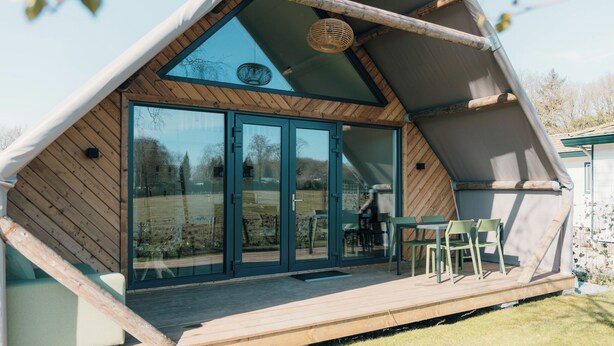 A modern, triangular-shaped building with large glass doors and a wooden deck with outdoor seating. - Big lodge | 6 persons (Voorthuizen)
