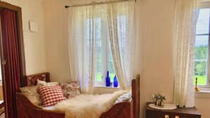 A bedroom with a wooden bed, white curtains, and a window with a view of greenery.