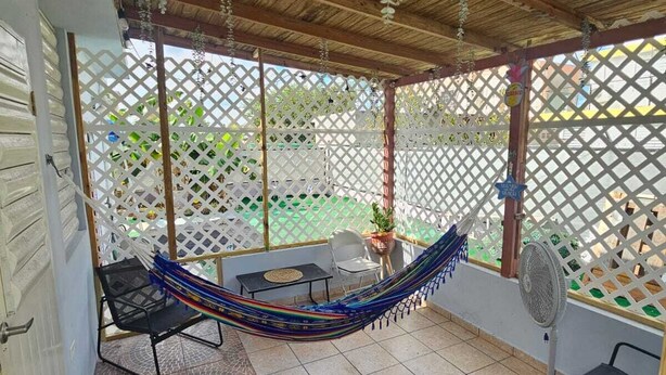A covered patio with a hammock, two chairs, a table, and a fan.