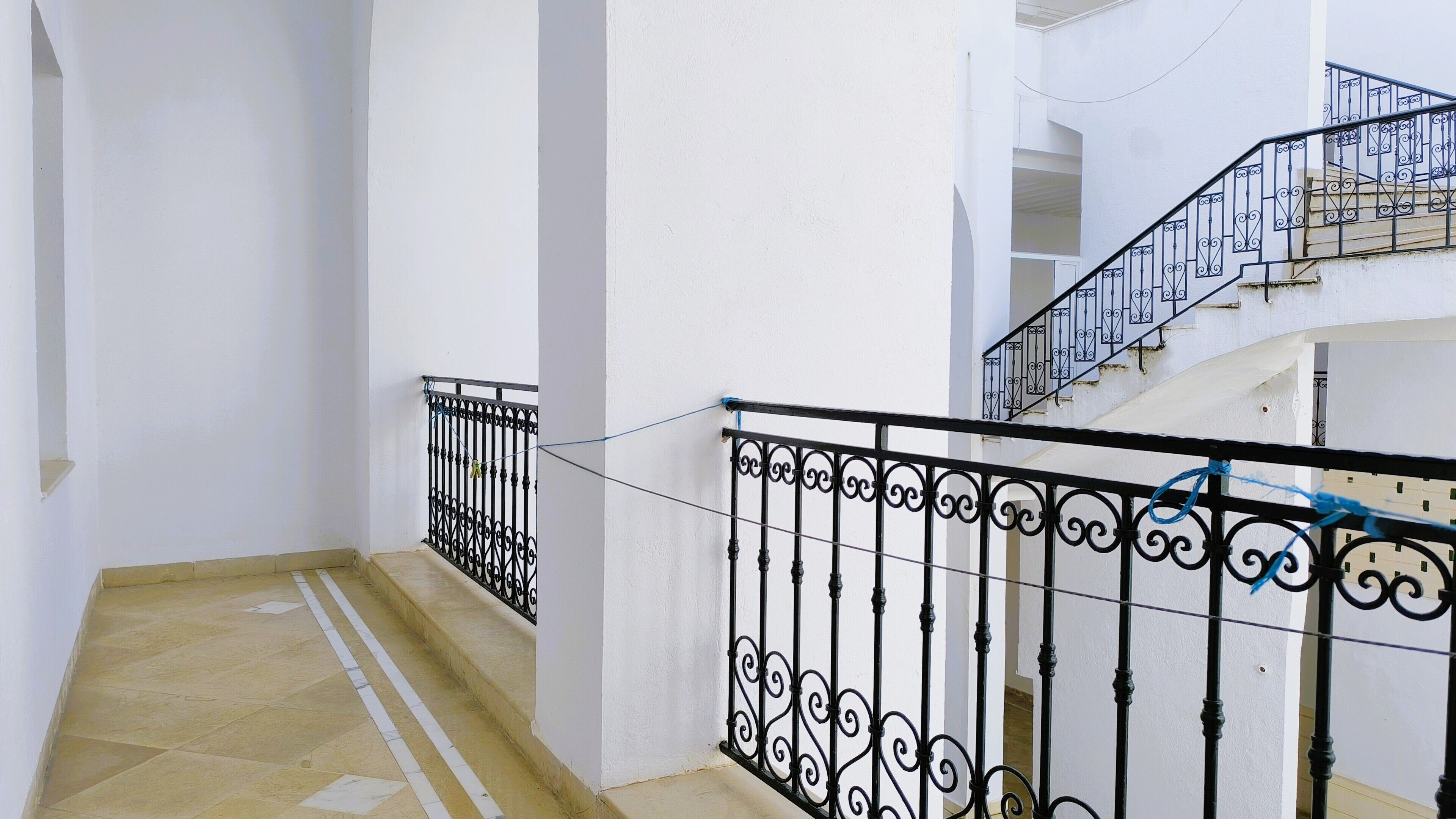 Un couloir aux murs blancs et au sol carrelé, qui mène à un escalier doté de rampes en fer forgé noir.