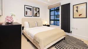 A bedroom with a bed, a nightstand, a window with curtains, and framed artwork on the wall. - The Melbray Gem - 2bdr Apartment With Courtyard (London)