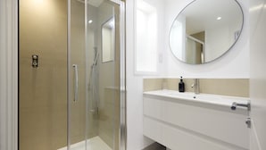 A modern bathroom with a glass-enclosed shower, a white vanity with a round mirror, and a beige tiled wall. - The Melbray Gem - 2bdr Apartment With Courtyard (London)