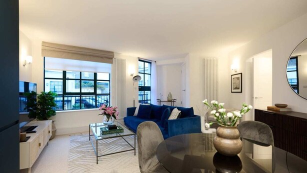 A modern living room with a blue sofa, a glass coffee table, and a dining table with chairs. There are large windows with curtains, a potted plant, and a mirror on the wall. - The Melbray Escape - Trendy 2bdr Apartment (London)