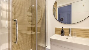 A modern bathroom with a glass-enclosed shower, a round mirror, and a white vanity with a wooden panel backsplash. - The Melbray Escape - Trendy 2bdr Apartment (London)