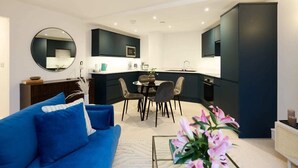 A modern kitchen with dark cabinetry, a round dining table with chairs, and a blue sofa in the living area. - The Melbray Escape - Trendy 2bdr Apartment (London)