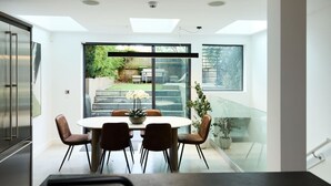 Un'area pranzo moderna con un tavolo rotondo e sedie, vista su un giardino attraverso ampie finestre, e una scala che conduce all'esterno.