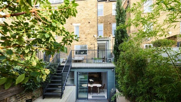 Un appartamento moderno con ingresso tramite porta in vetro, un piccolo spazio esterno con tavolo e sedie, e una scala che conduce a un balcone.